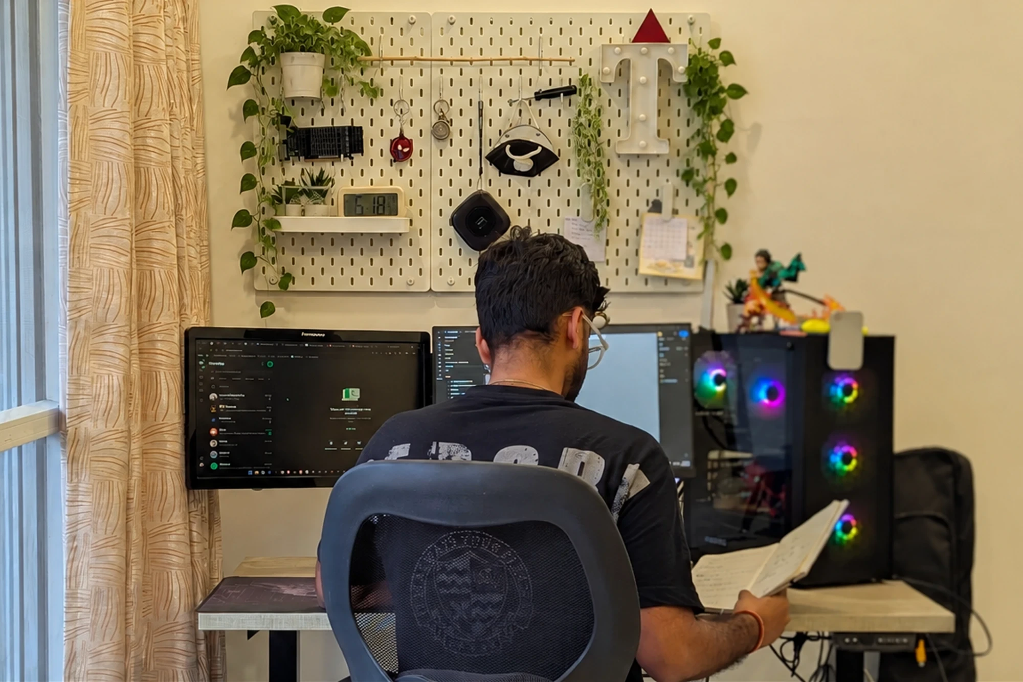 Tanishq at his desk, working across two monitors, with a pegboard of plants, a clock and a light-up T on the wall above